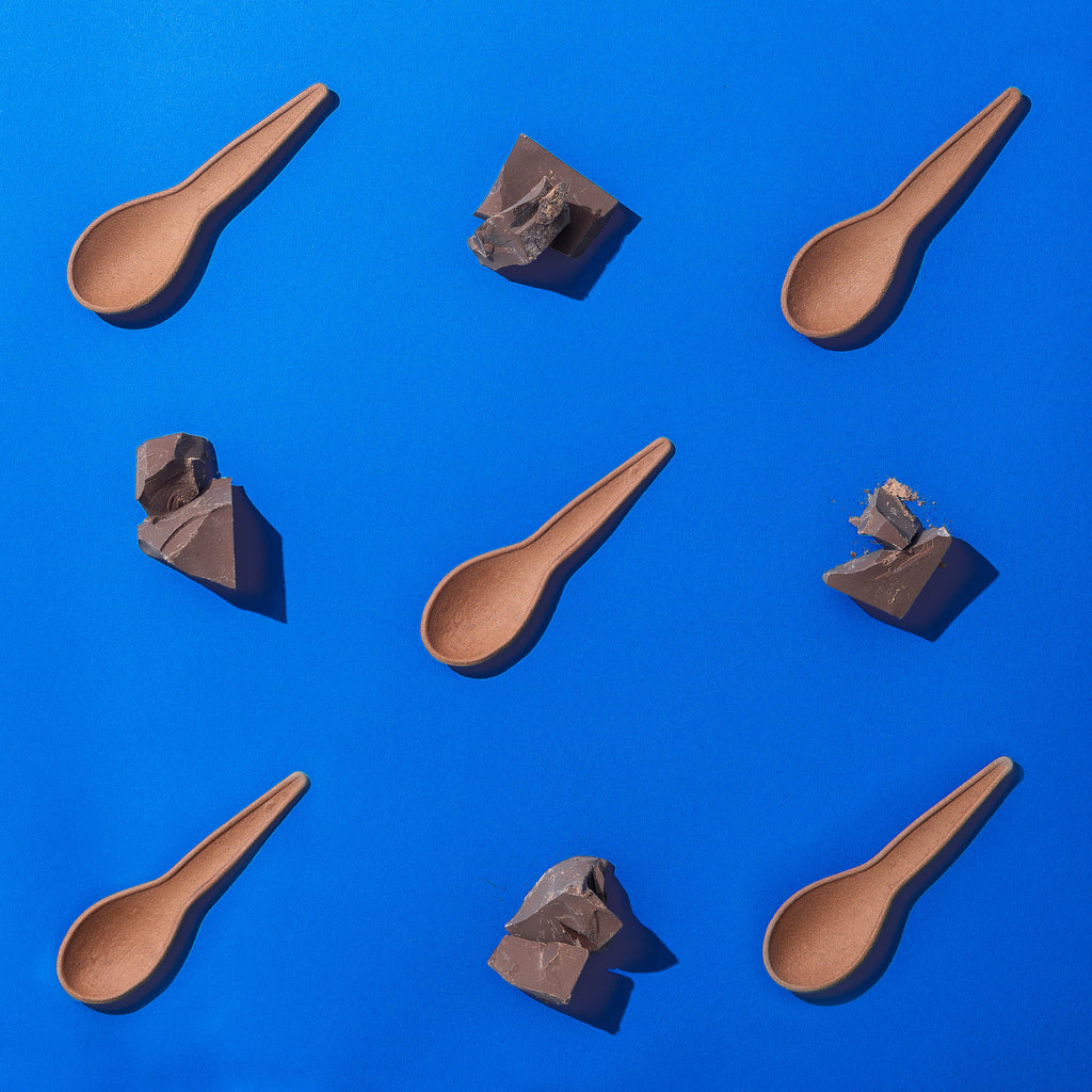 A photo of chocolate edible spoons laid out in a pattern with chunks of raw chocolate. The background is blue.