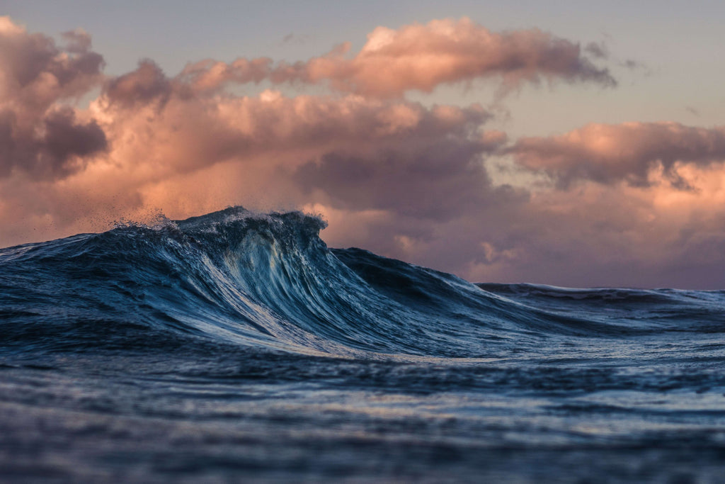 A photo of an ocean wave. There is a pink and purple sunset sky and clouds in the background.